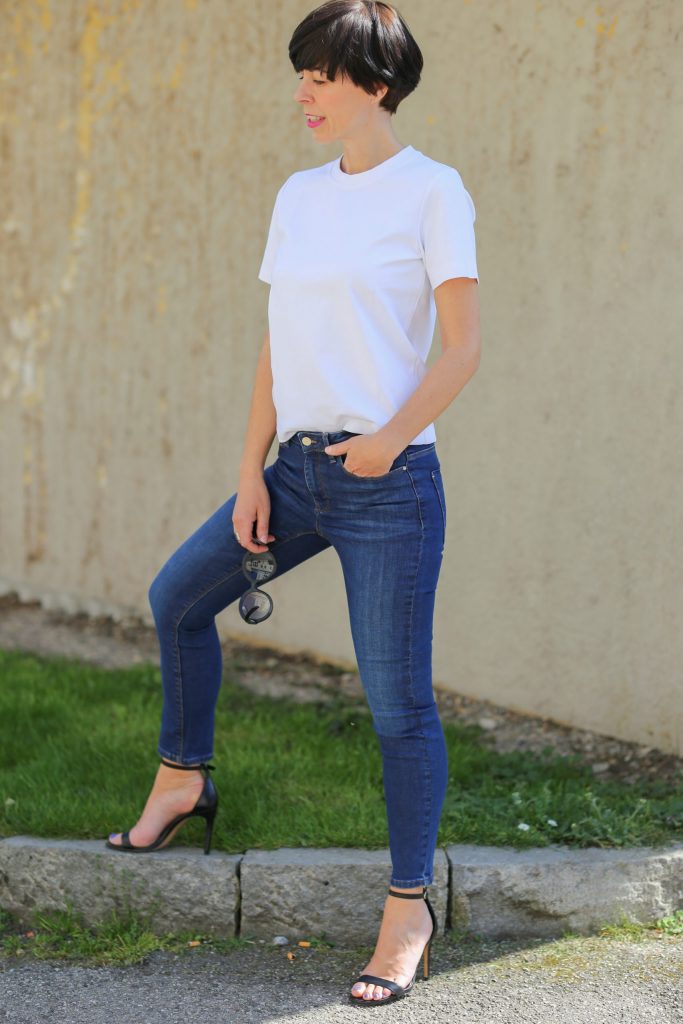 nachhaltige jeans Frau mit kurzem schwarzem Haar und schwarzer Sonnenbrille in der Hand trägt ein weisses T-Shirt und Skinny Hosen