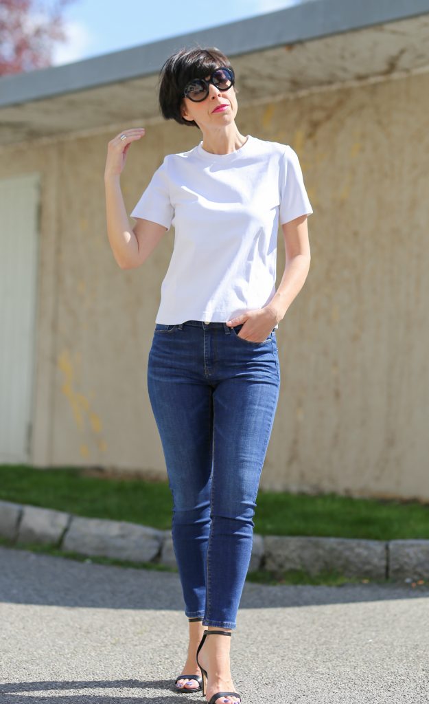 nachhaltige jeans Frau mit kurzem schwarzem Haar und schwarzer Sonnenbrille trägt ein weisses T-Shirt und Skinny Hosen und High Heels